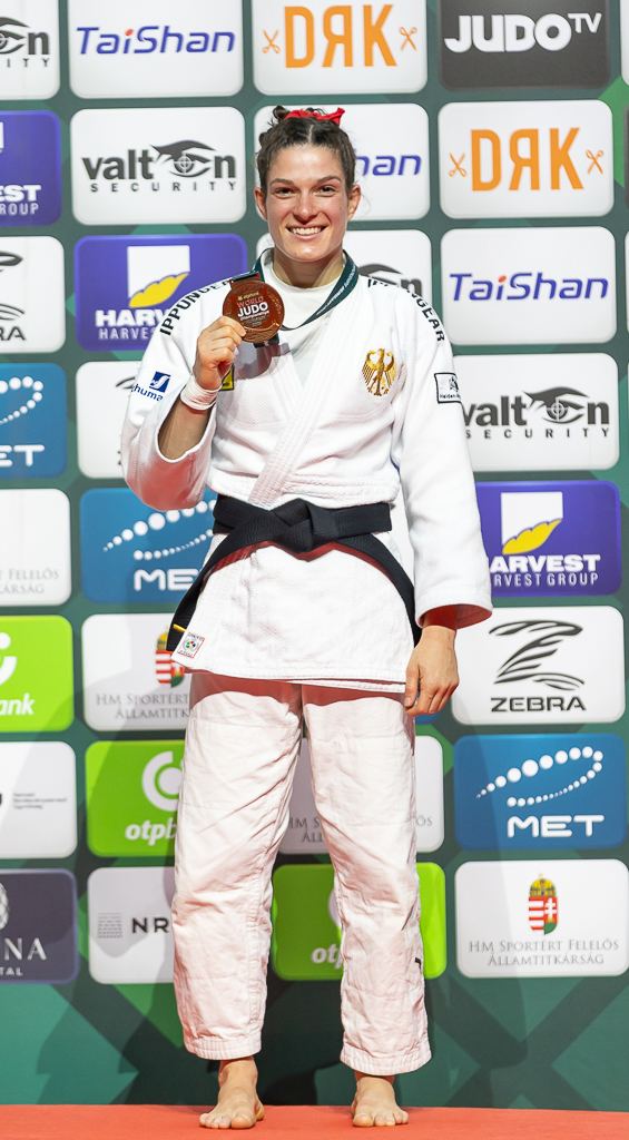 Mascha steht im Judoanzug mit der Bronzemedaille in der Hand vor einer Werbewand und lächelt strahlend in die Kamera.