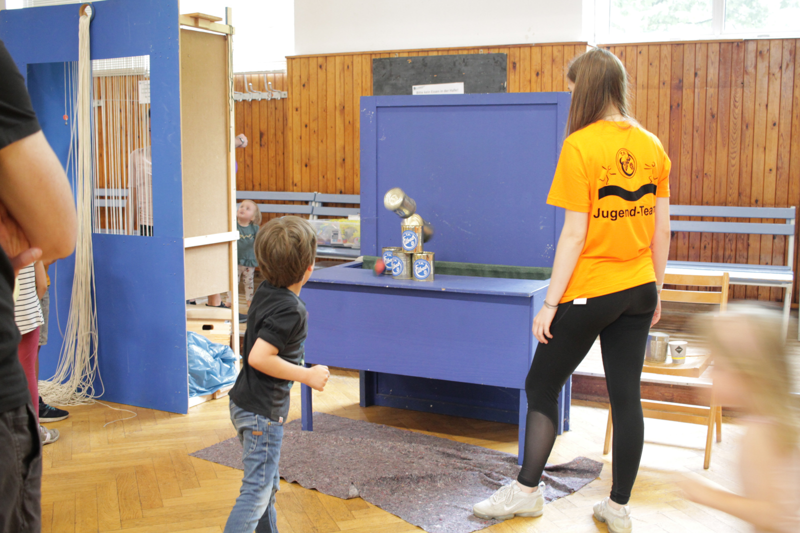 Ein Kind in Jeans uns schwarzem T-Shirt am Dosenwerfen-Stand. Der Ball trifft den Dosenstapel. Daneben steht eine Helferin des THE-Jugendausschuss im orangenen Jugend-T-Shirt.