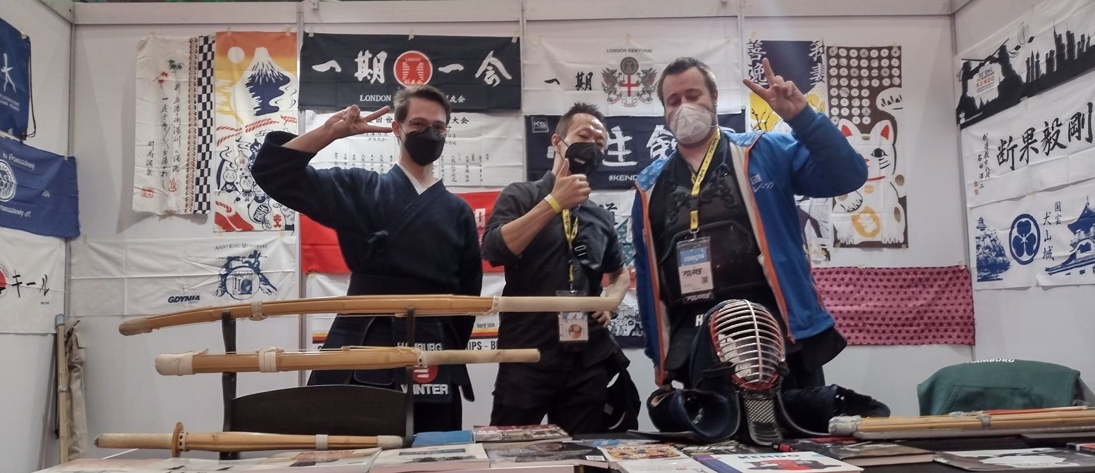 Drei Männer mit Mund-Nasen-Schutz stehen hinter einem Messetresen und machen das "Peace"-Zeichen. Im Vordergrund sind einige japanische Schwerter zu sehen und im Hintergrund die Rückwand des Messe-Stands mit viele Plakate und Bildern.
